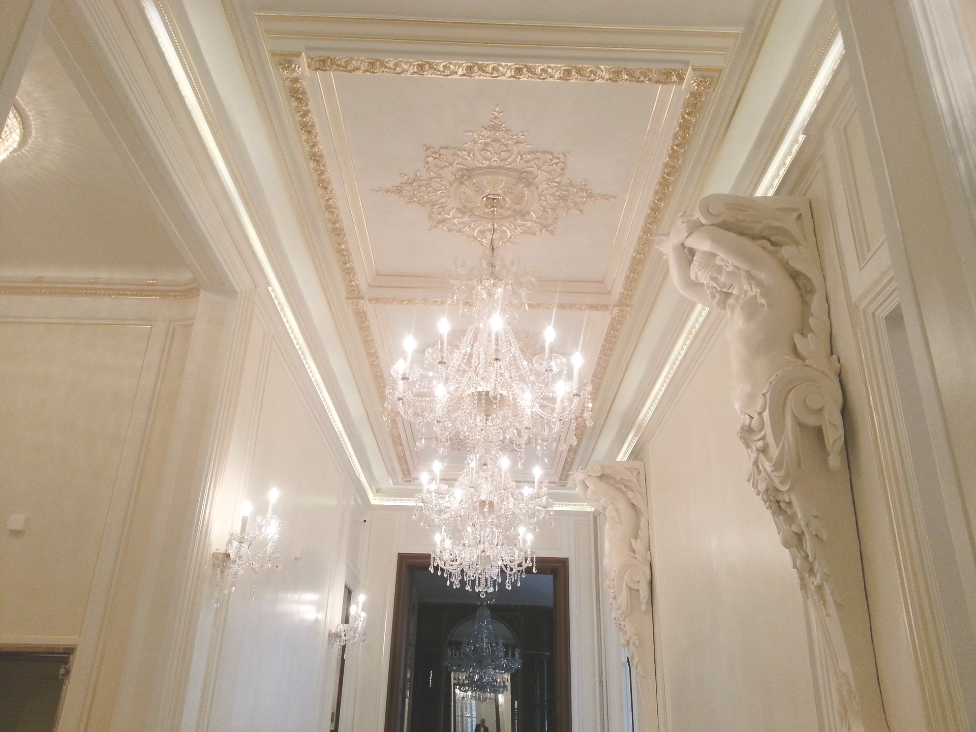 Un grand couloir couleur crème avec un lustre en cristal, des moulures de plafond ornementées et des supports muraux.
