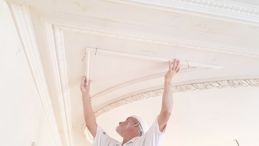Une personne en uniforme blanc installe une moulure décorative sur un plafond blanc.
