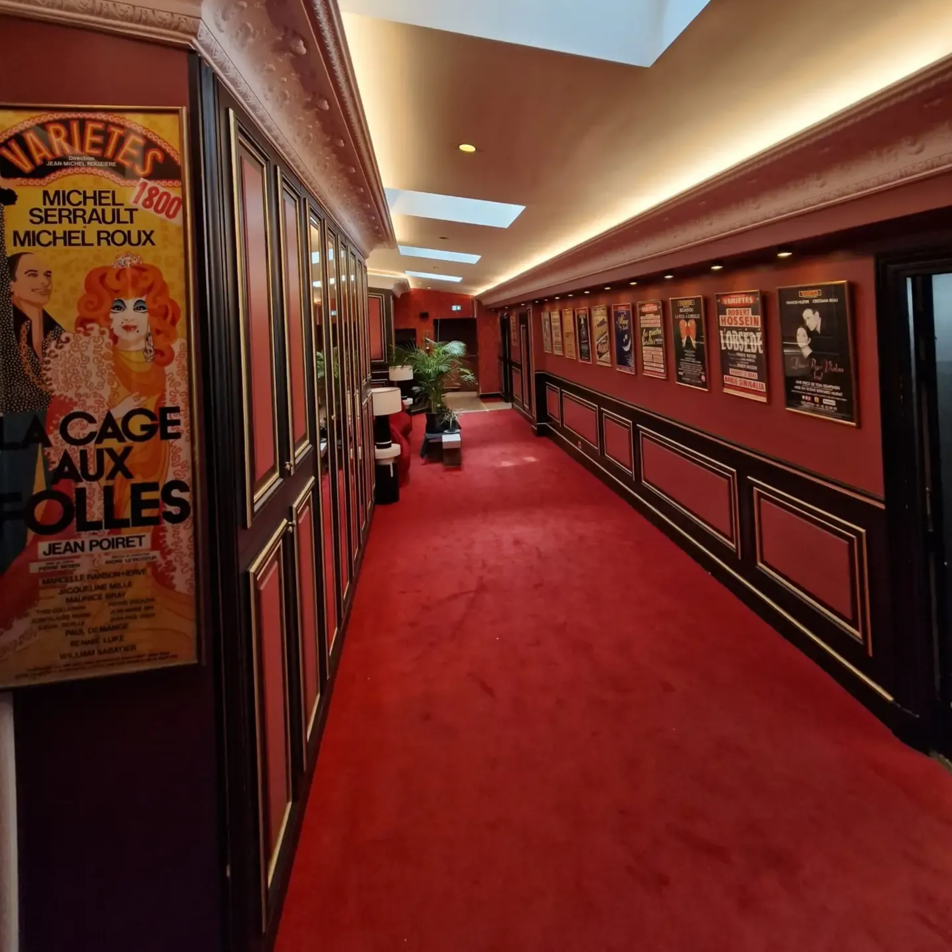 Un couloir recouvert de moquette dans un théâtre, bordé d'affiches de films et de pièces de théâtre sur des murs rouge.