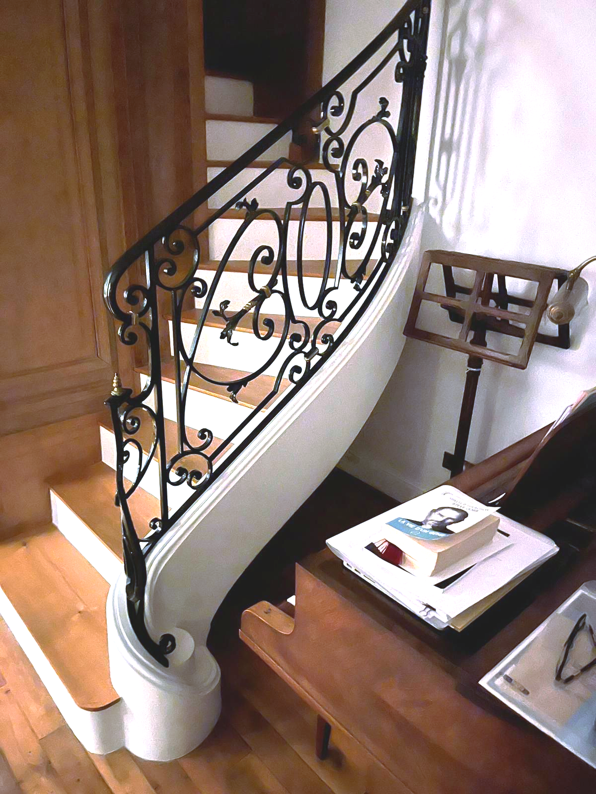 Un escalier en bois avec une rampe en fer noir ouvragée, à côté d'un piano sur lequel se trouvent des livres.