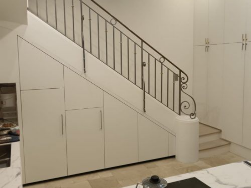 Des meubles de rangement blancs ont été intégrés dans l'espace sous un escalier doté d'une rampe décorative en métal foncé.