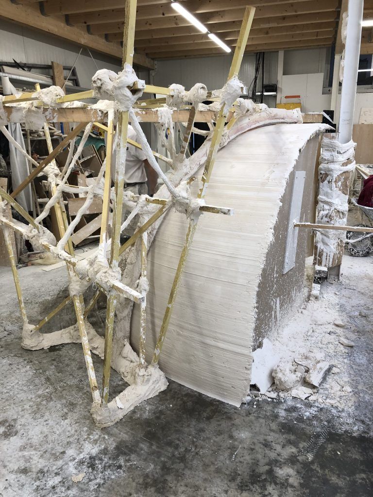 Dans un atelier, une structure de sculpture incurvée partiellement construite, soutenue par des poutres en bois.