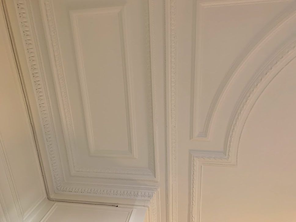 Moulure décorative blanche au plafond, avec des panneaux rectangulaires et des détails architecturaux arrondis.