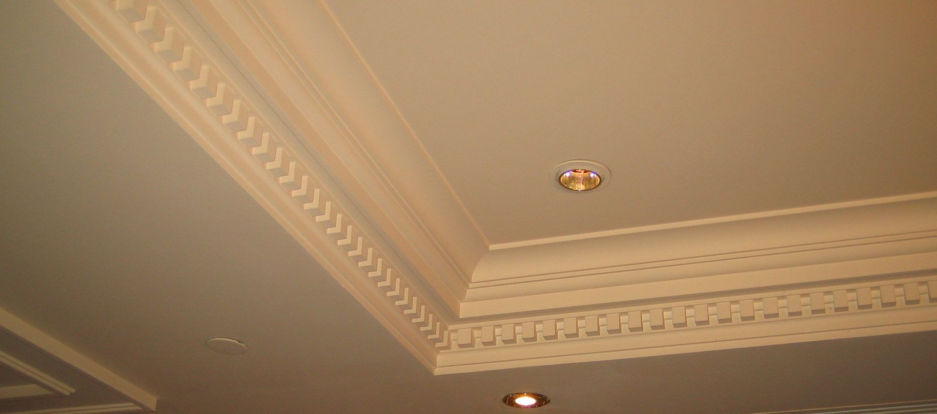 Un angle de plafond orné d'une moulure de corniche couleur crème aux détails complexes et de deux spots encastrés.