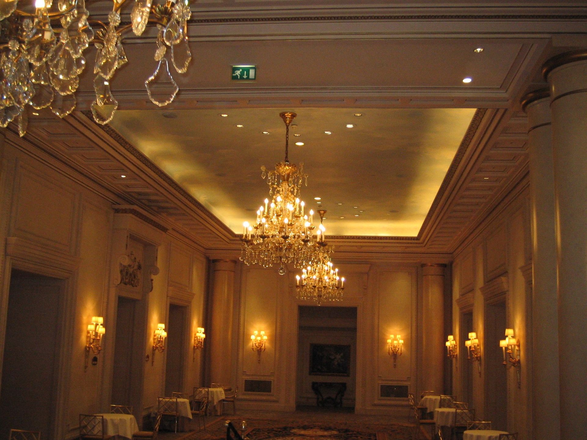 Une pièce spacieuse et faiblement éclairée, ornée de lustres somptueux, de hautes colonnes et de plusieurs tables.