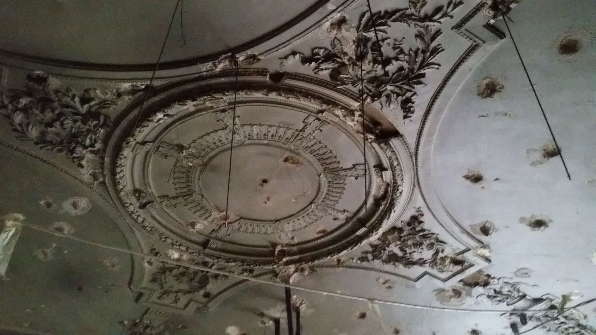 Moulures de plafond ornementées, avec des décors floraux en plâtre, abîmées et écaillées, dans une pièce sombre et délabrée.