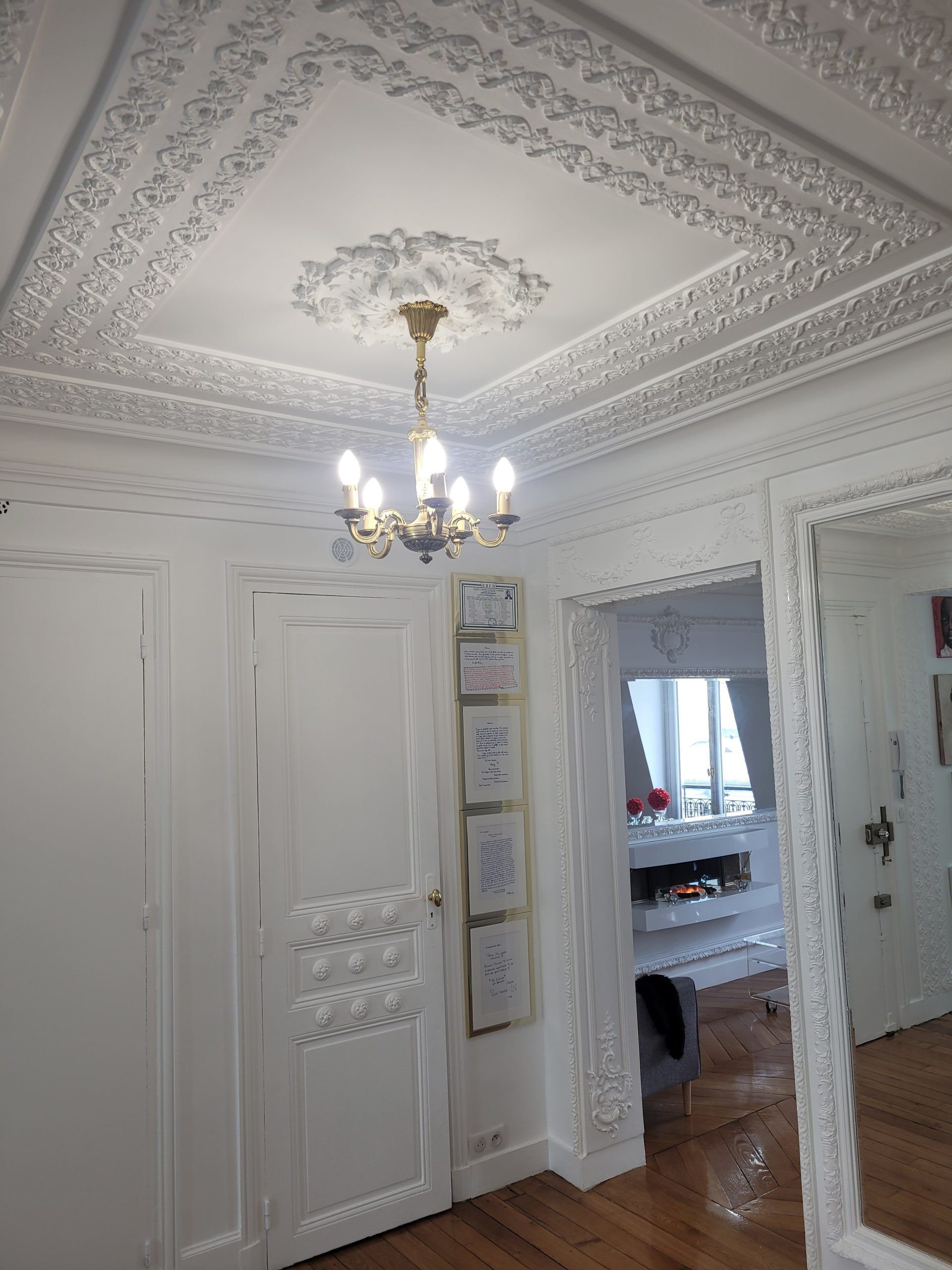 Un couloir orné, peint en blanc, avec des moulures de plafond détaillées, un lustre ancien et un grand miroir décoratif.