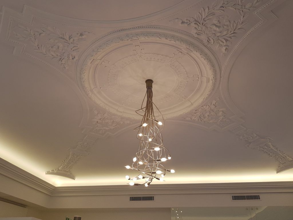 Dans une pièce aux tons blancs, un lustre en cristal à plusieurs branches est suspendu à un médaillon.