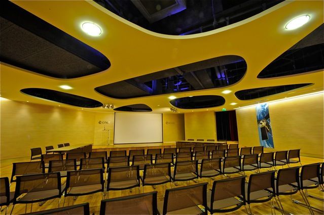 Une salle de séminaire moderne avec un écran de projection sous un plafond orné de formes découpées.