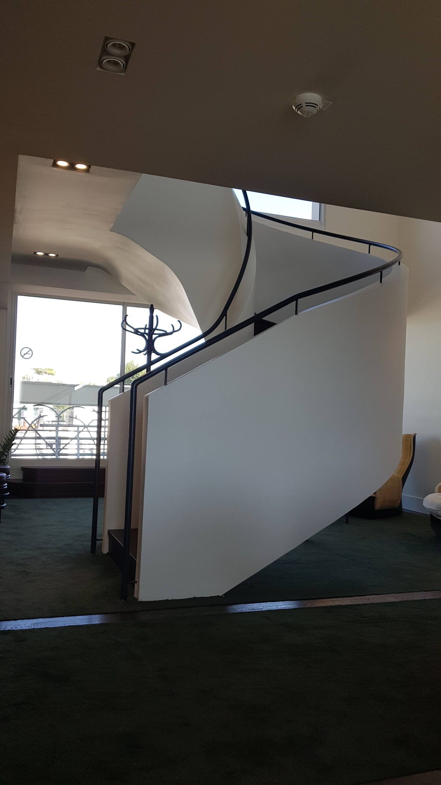 Un escalier blanc en colimaçon se dresse dans une pièce baignée de soleil, au sol recouvert de moquette.