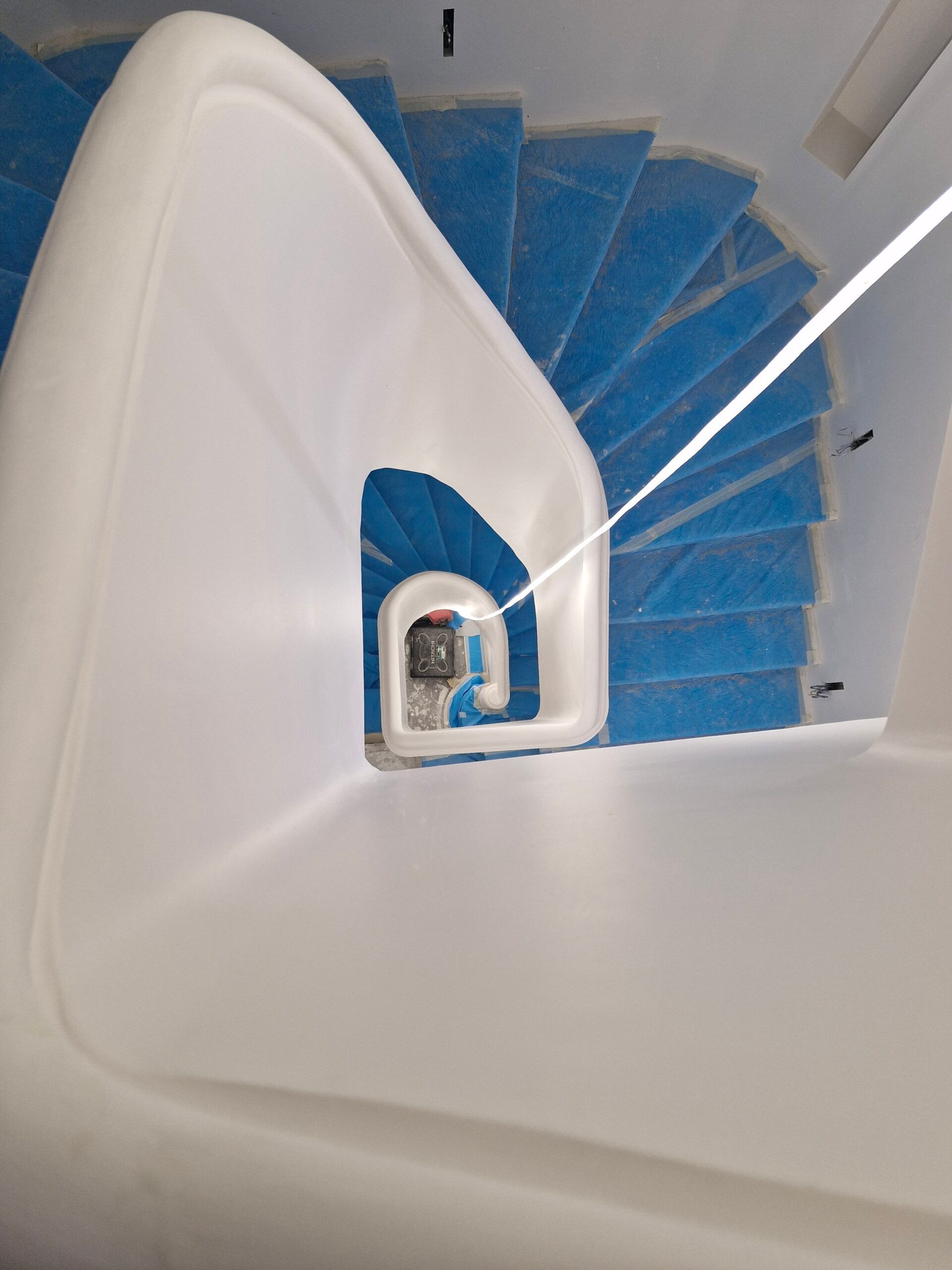 Vue plongeante sur un escalier en colimaçon aux rampes blanches et film protecteur bleu recouvrant chaque marche.
