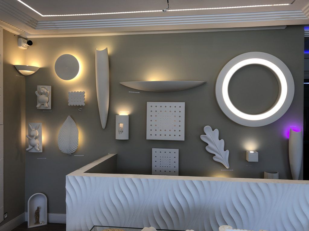 Un mur d'exposition présentant des appliques murales, géométriques et organiques, éclairées par une lumière LED.