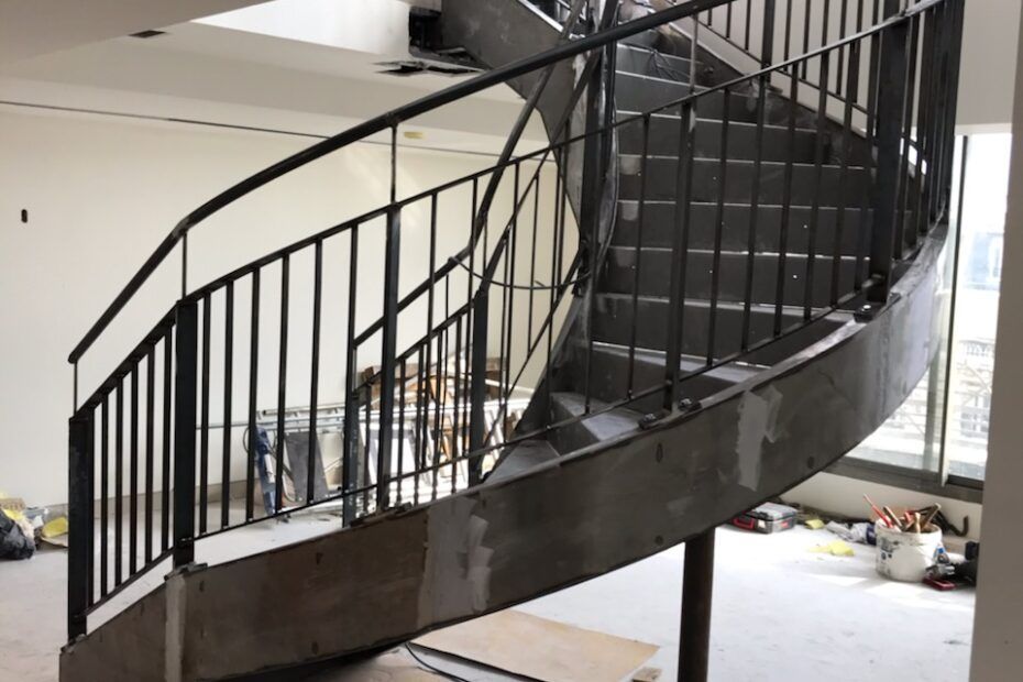 Un escalier métallique incurvé en construction dans une pièce inachevée, avec des rampes en métal gris et une base en béton.