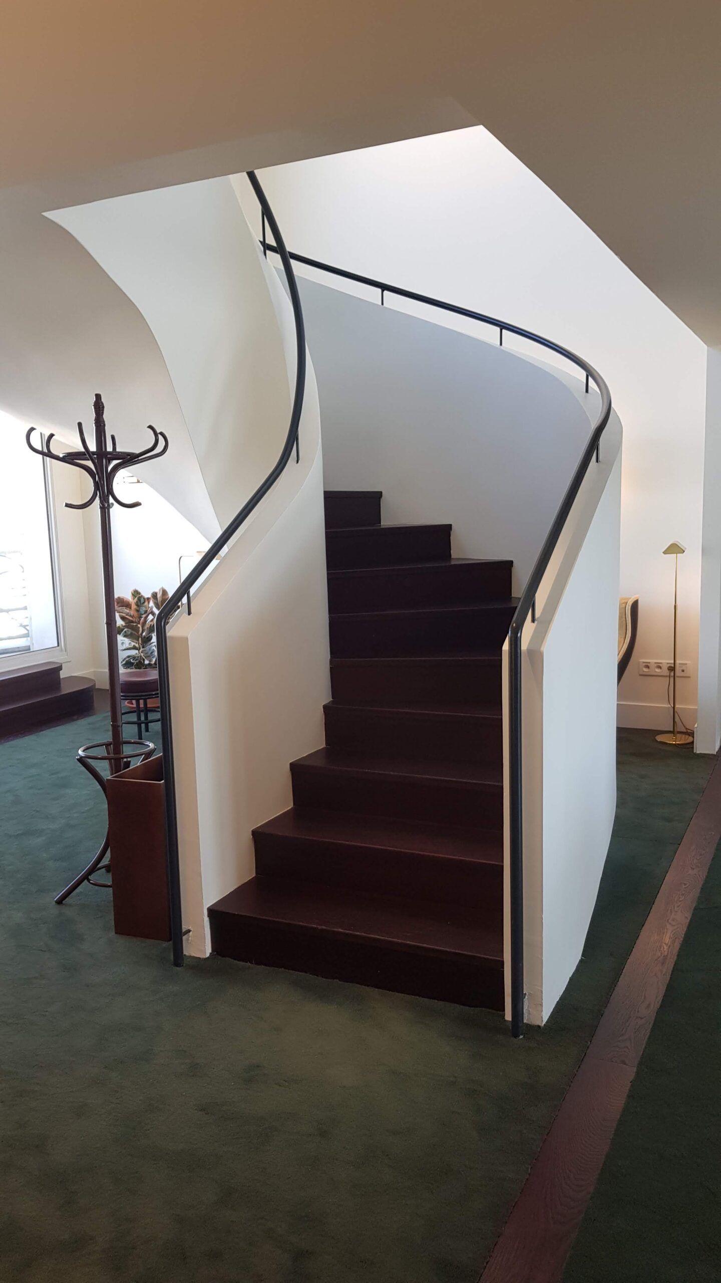 Un escalier courbe aux marches en bois foncé et aux murs blancs, avec une rampe en métal et un porte-manteau au premier plan.