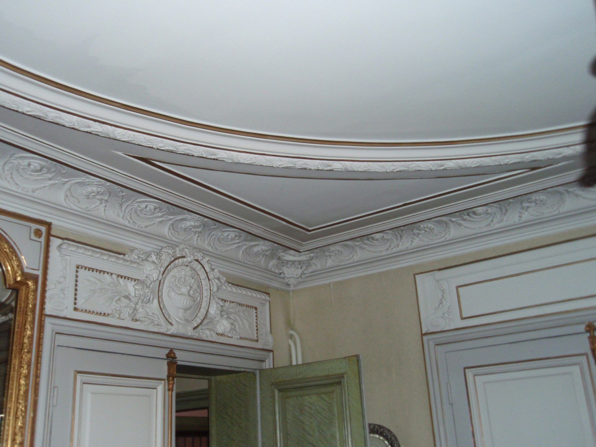 Une moulure ornementale blanche et dorée décore le coin d'une pièce aux murs et aux portes clairs.