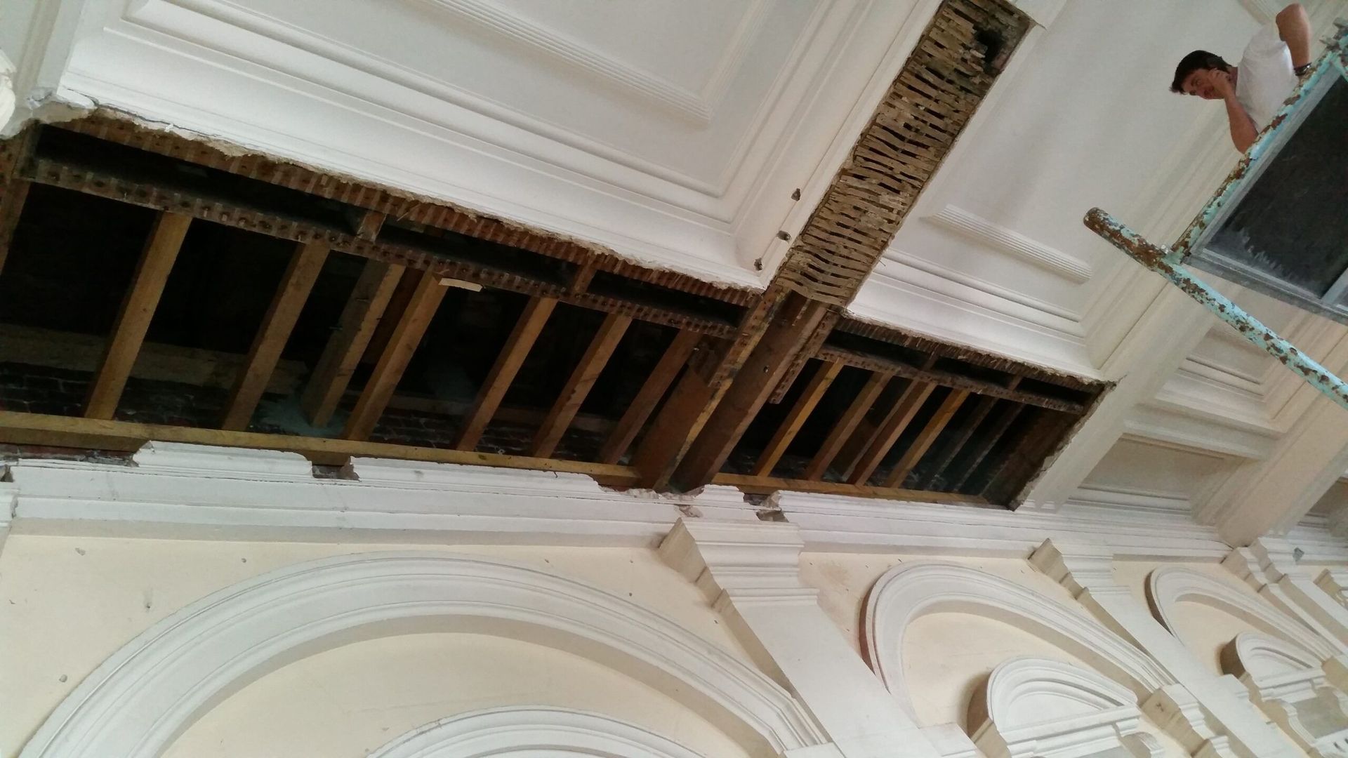 Un ouvrier, juché sur une échelle, répare un plafond orné endommagé, laissant apparaître les solives en bois.