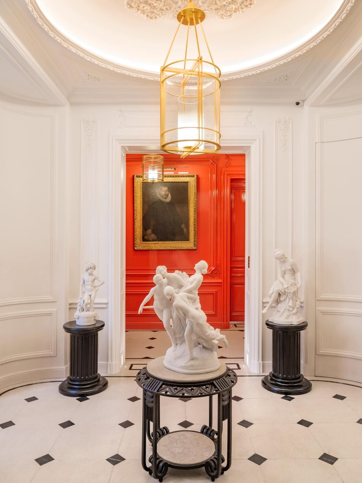 Un hall d'entrée élégant avec un plafond circulaire blanc, un luminaire suspendu doré, un sol carrelé, des statues blanches.