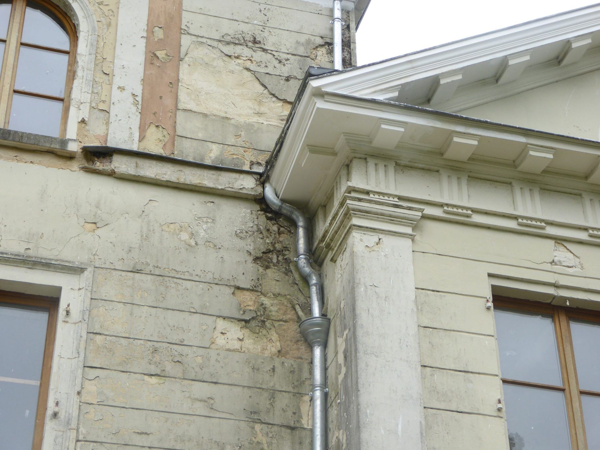 Un vieux bâtiment d'angle avec des murs couleur crème, des fenêtres et une gouttière métallique sous l'avant-toit.