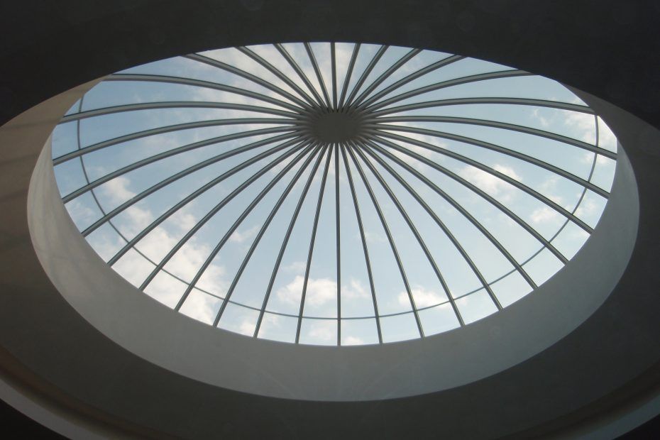 Une verrière circulaire à motif de grille rayonnante, donnant sur un ciel bleu parsemé de doux nuages ​​blancs.