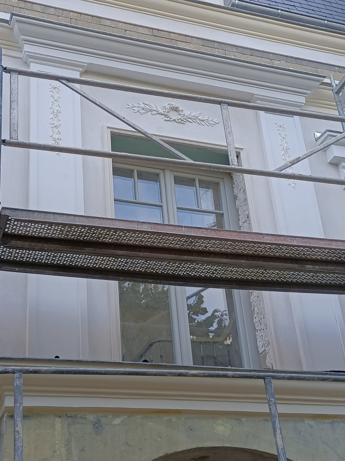 La façade d'un bâtiment de couleur claire, ornée d'une haute fenêtre à plusieurs carreaux et de moulures décoratives.