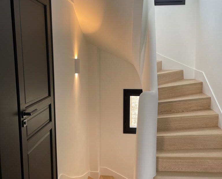 Un escalier intérieur moderne et lumineux avec des marches en bois, un mur blanc incurvé, une porte sombre.