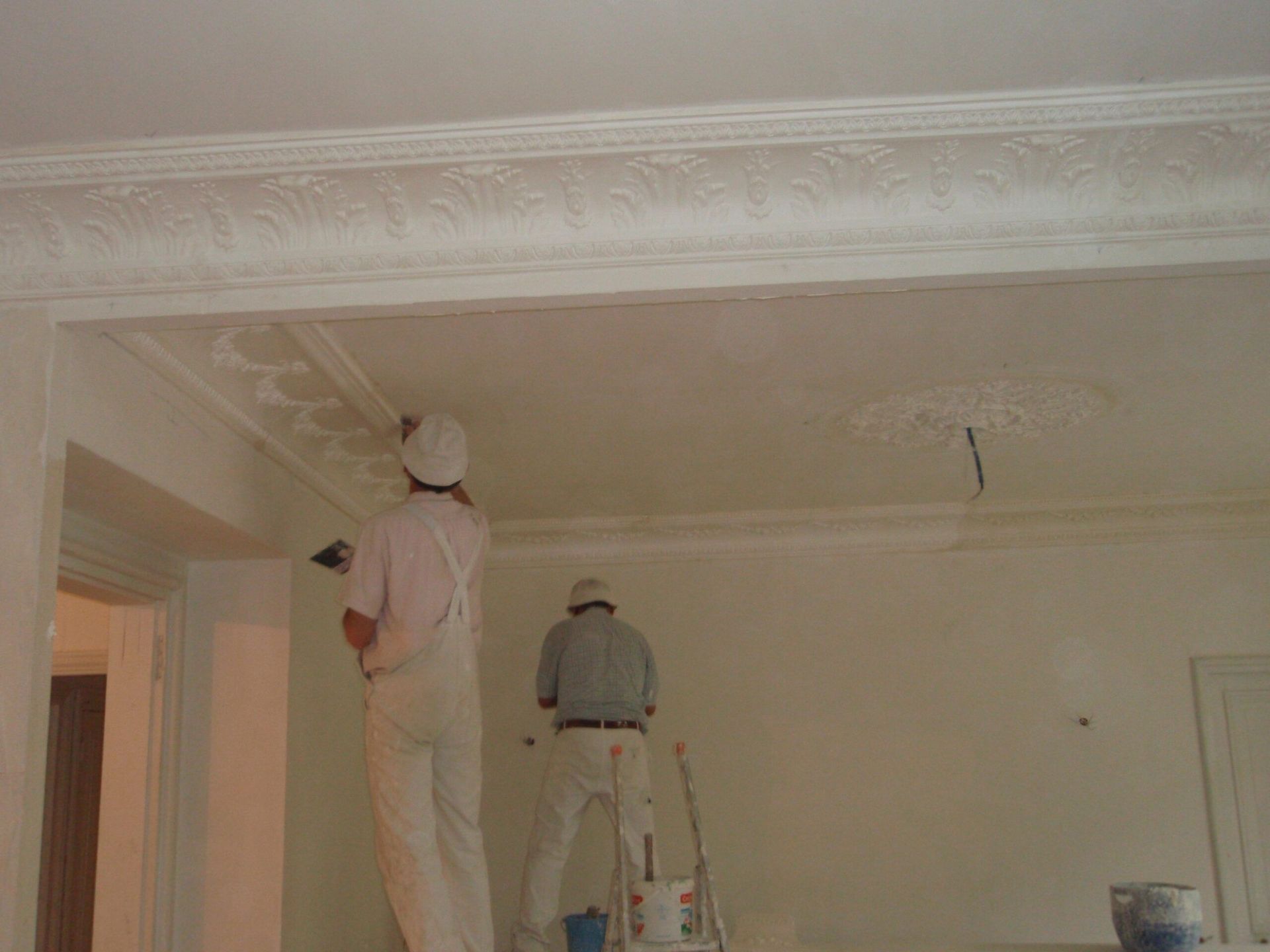 Deux ouvriers en salopettes blanches et casques de chantier appliquent du plâtre.