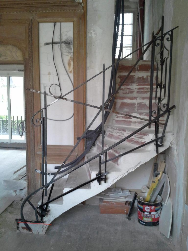 Un escalier avec une rampe décorative en métal noir, des marches en briques apparentes et des outils de construction.