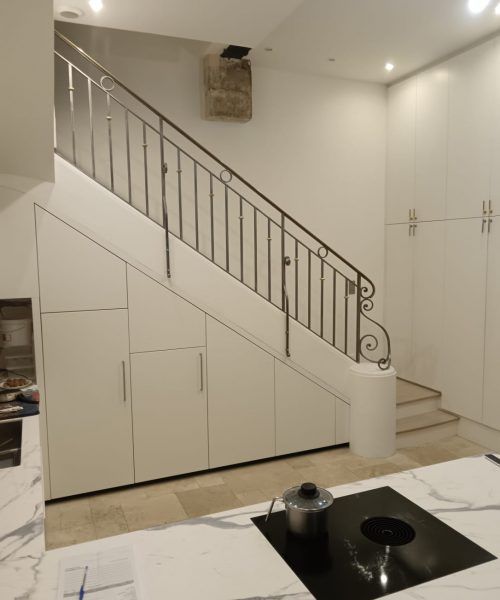 Des placards blancs sous un escalier, avec des poignées argentées, jouxtent un escalier avec une rampe en fer.