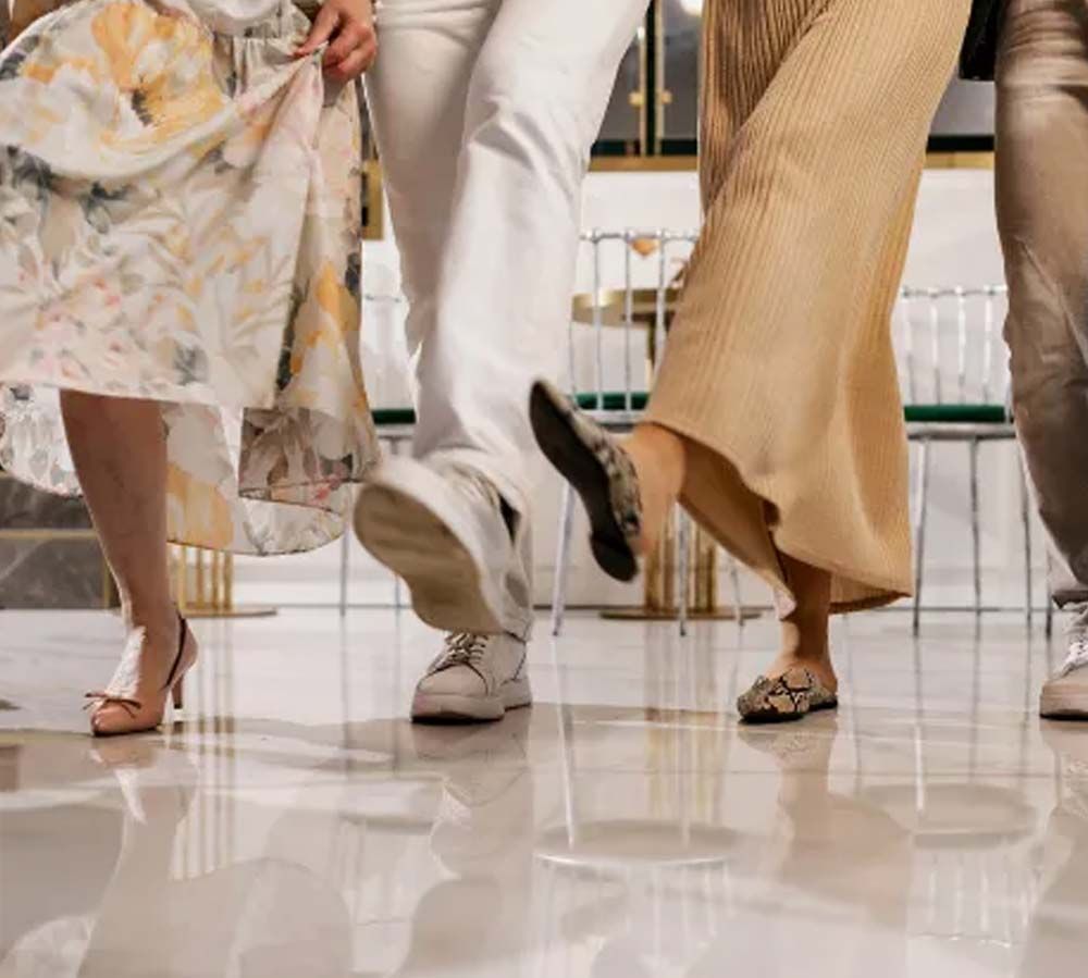 Tres personas caminando en el interior de un edificio, vistas desde las rodillas hacia abajo, vestidas con vestidos, pantalones y sandalias de colores claros.