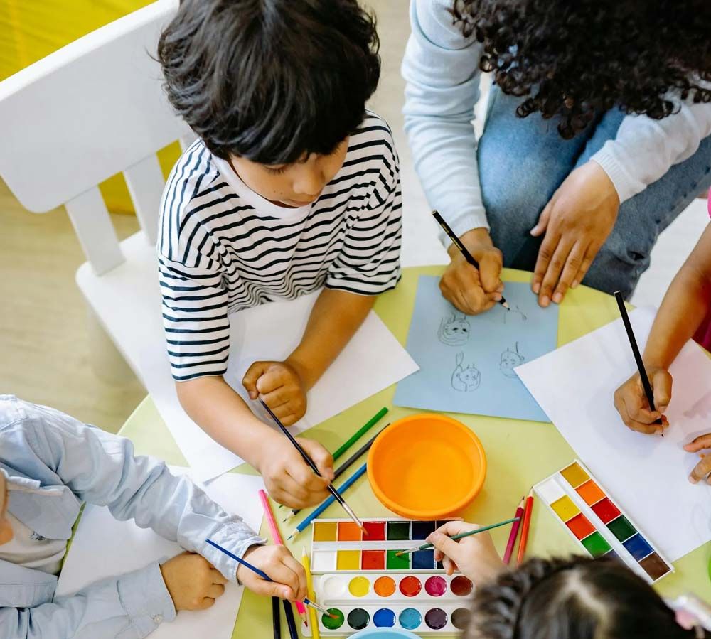 Niños dibujando y pintando juntos en una mesa con crayones, papel y un recipiente con pintura.