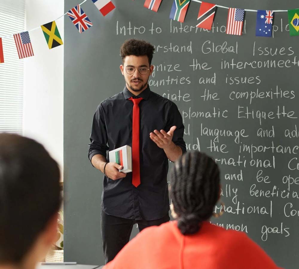 El profesor habla en el aula frente a la pizarra con banderas internacionales y notas escritas.