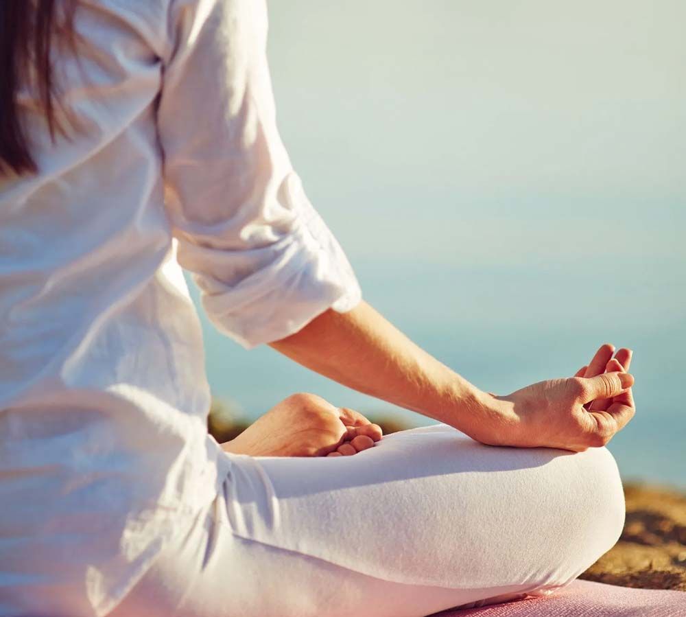 Persona vestida de blanco meditando con las piernas cruzadas junto al agua al atardecer.