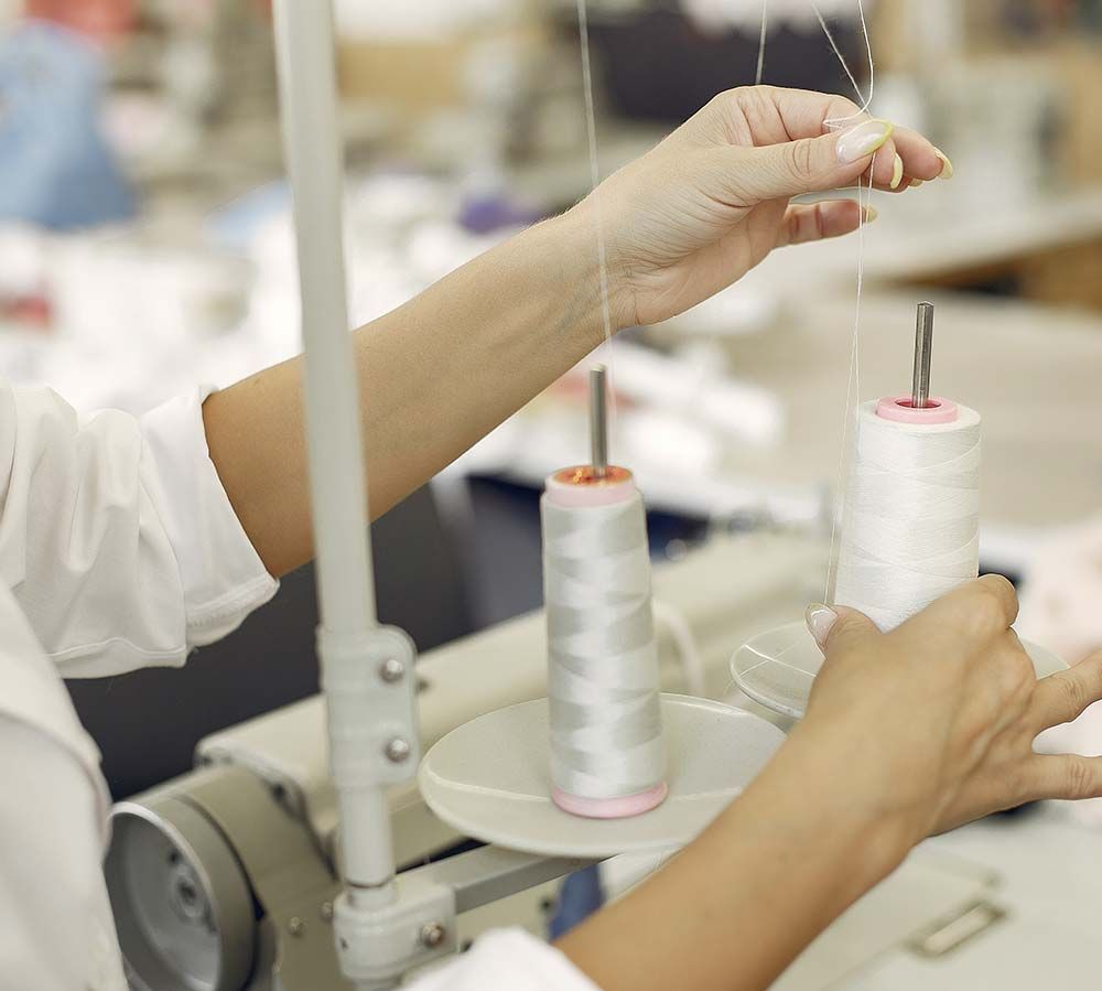 Manos trabajando con carretes de hilo blanco en una máquina de coser en un taller.