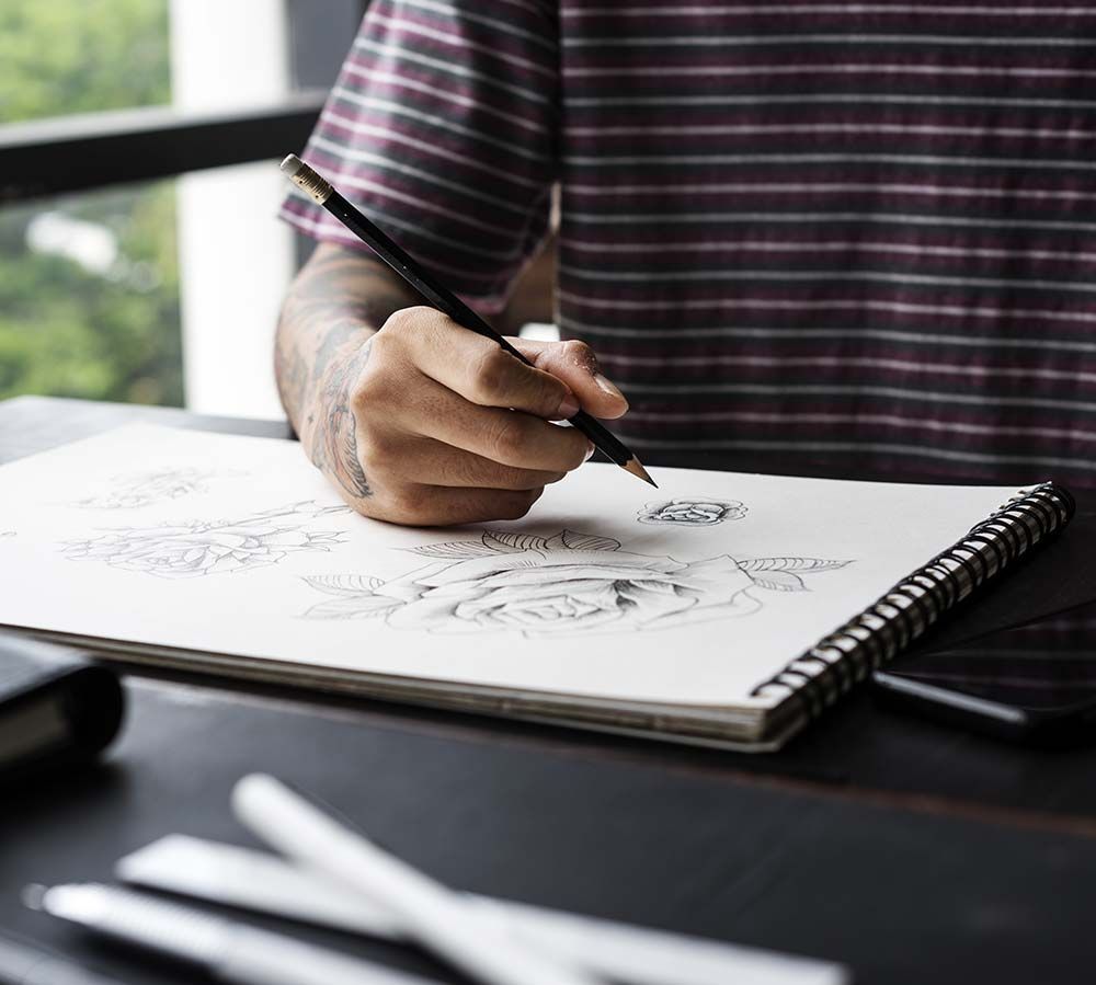 Una persona dibuja en un cuaderno de espiral sobre un escritorio con herramientas de arte cerca.