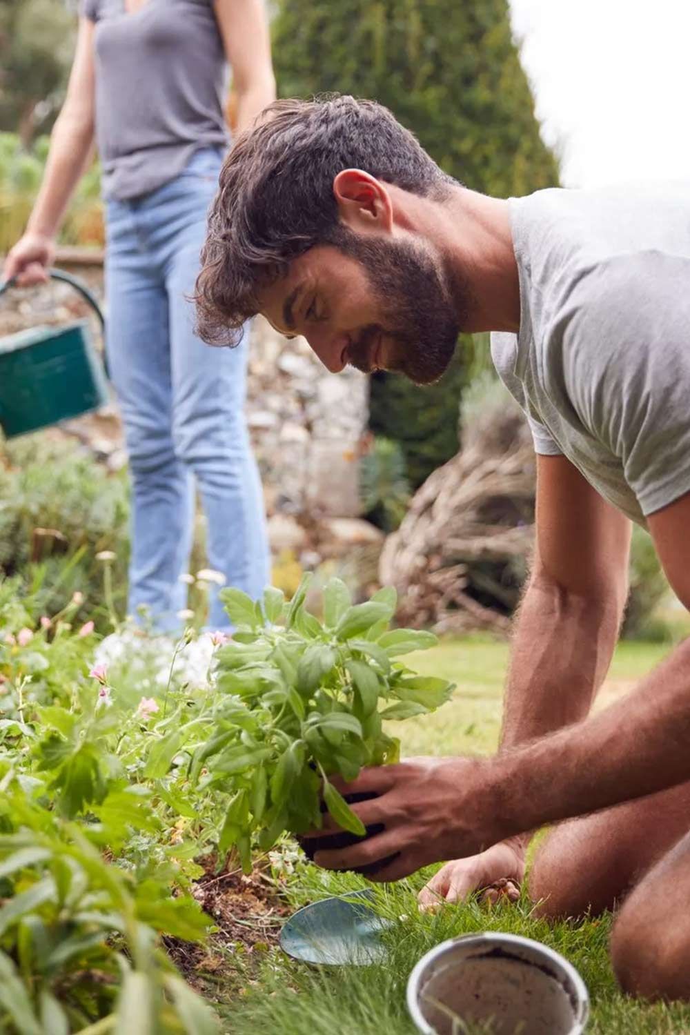 Un hombre se arrodilla para plantar una planta en un jardín mientras una mujer riega las plantas.