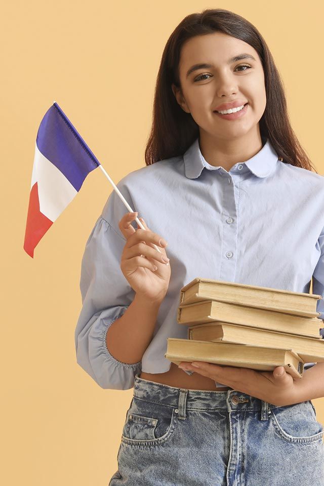 Estudiante sonriente sosteniendo una bandera francesa y una pila de libros sobre un fondo amarillo.