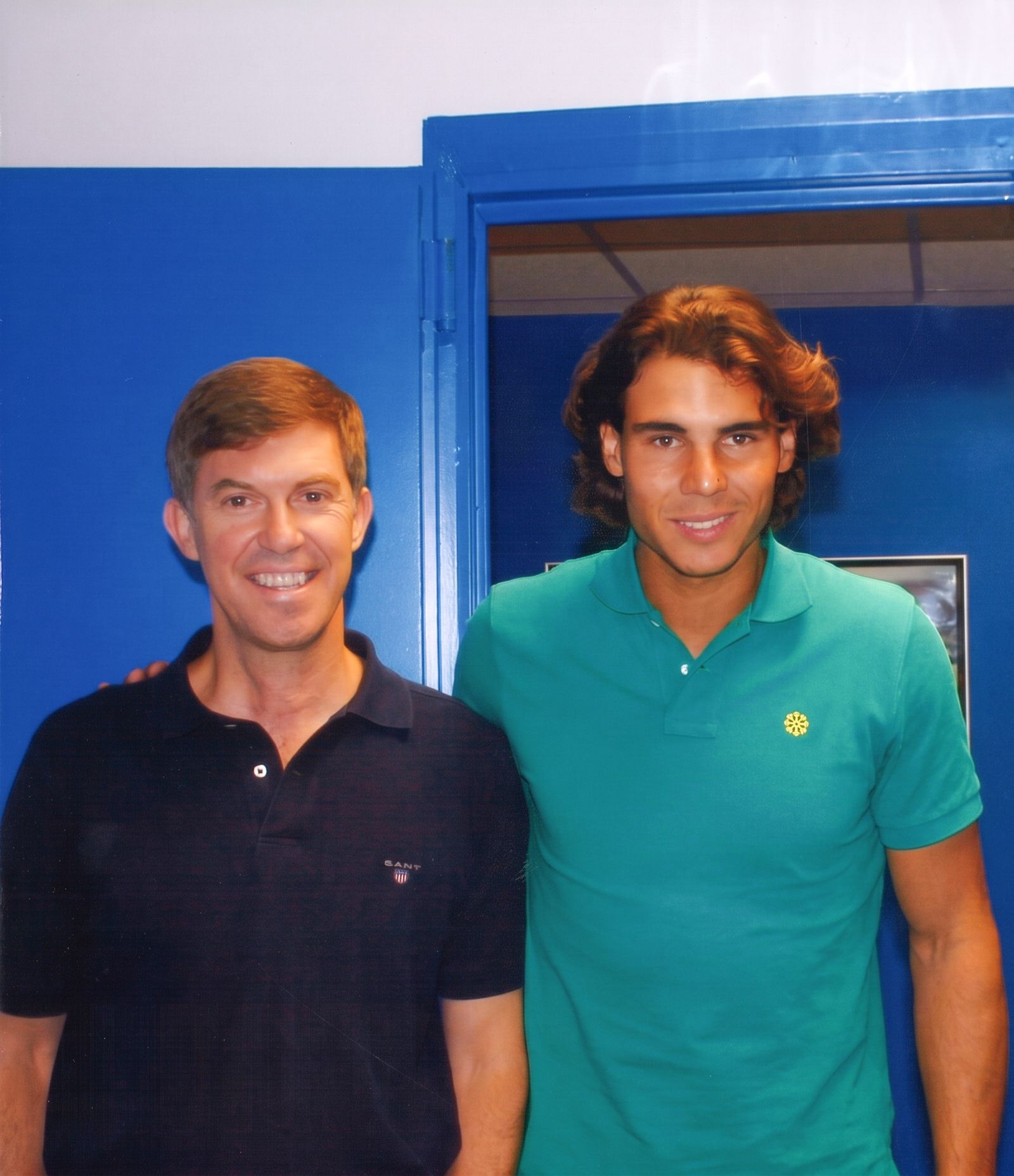 Dos hombres de pie en el interior, junto a una puerta azul; ambos sonríen. Uno lleva un polo oscuro y el otro, uno verde.