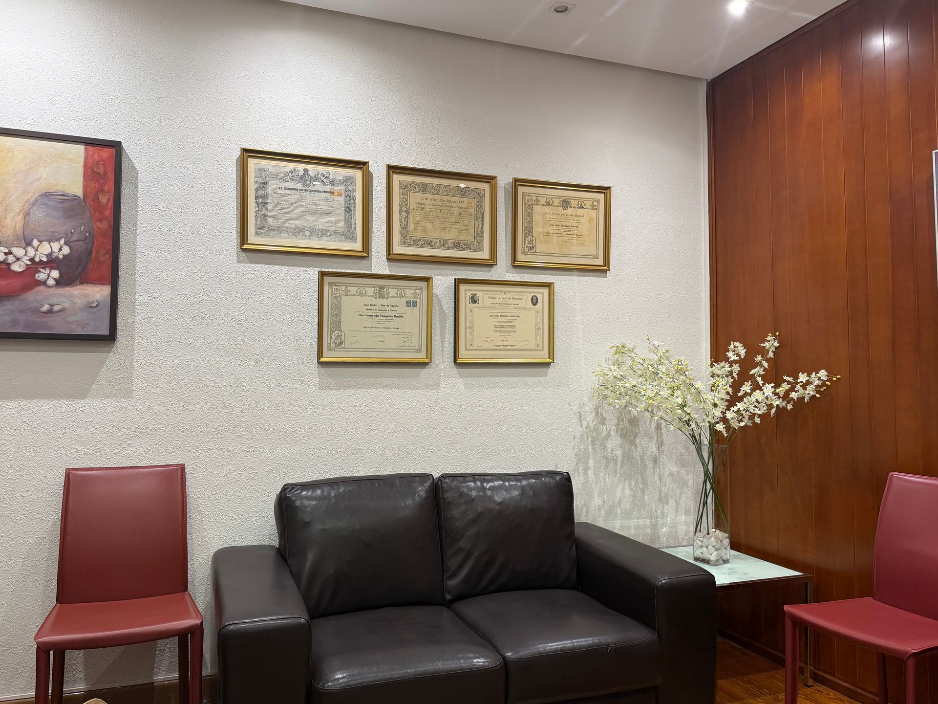 Zona de espera con sofá negro, sillas rojas, certificados enmarcados y un jarrón con flores blancas.
