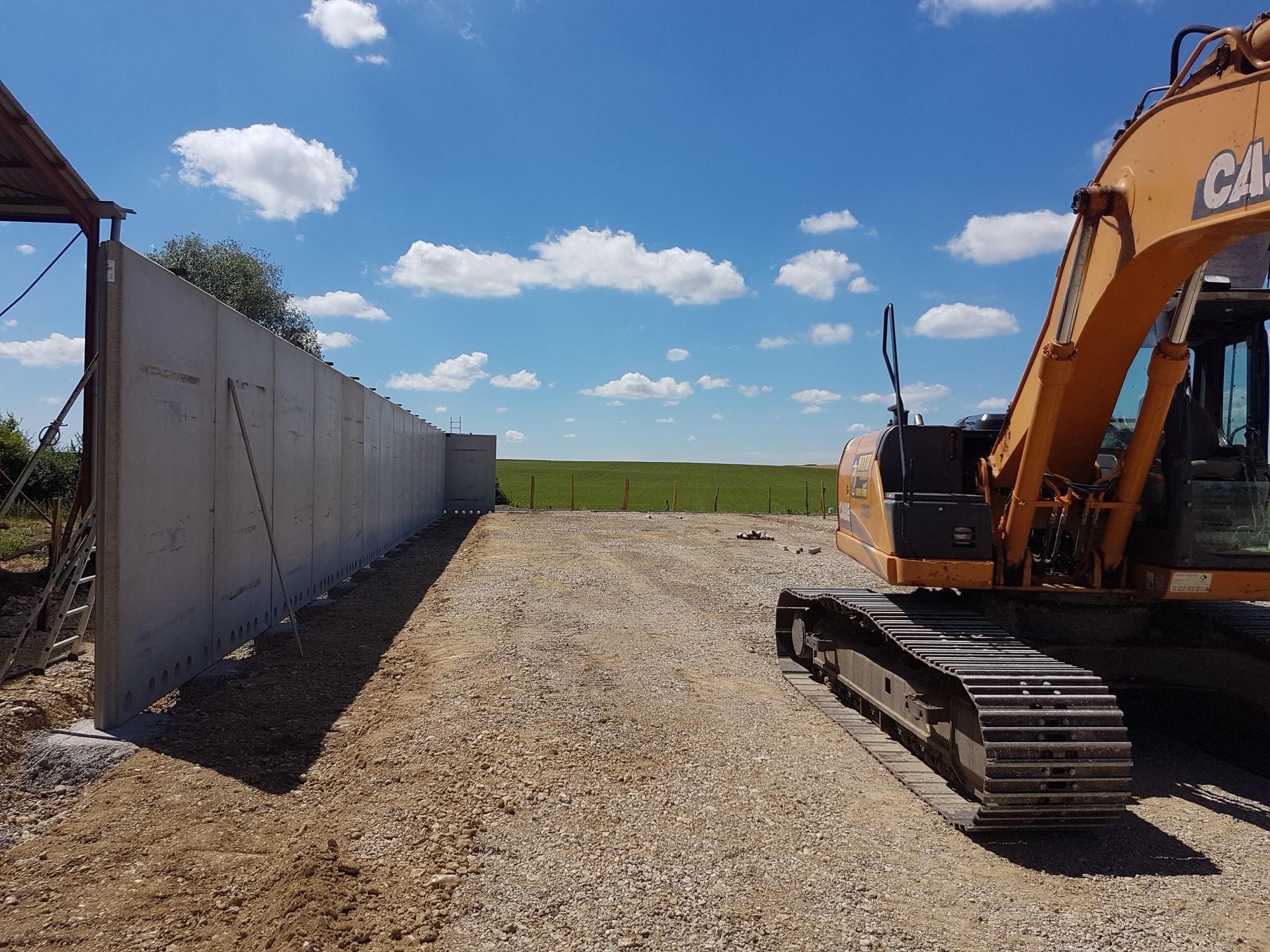 Chantier de construction avec une excavatrice sur du gravier à côté d'un mur en béton sous un ciel bleu.