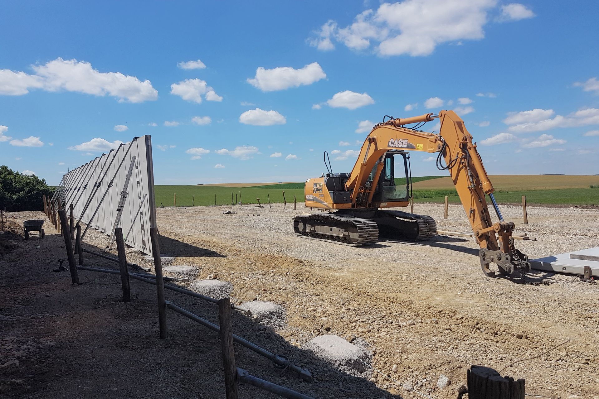 Une excavatrice travaille sur un chantier de construction en gravier sous un ciel bleu parsemé de nuages.