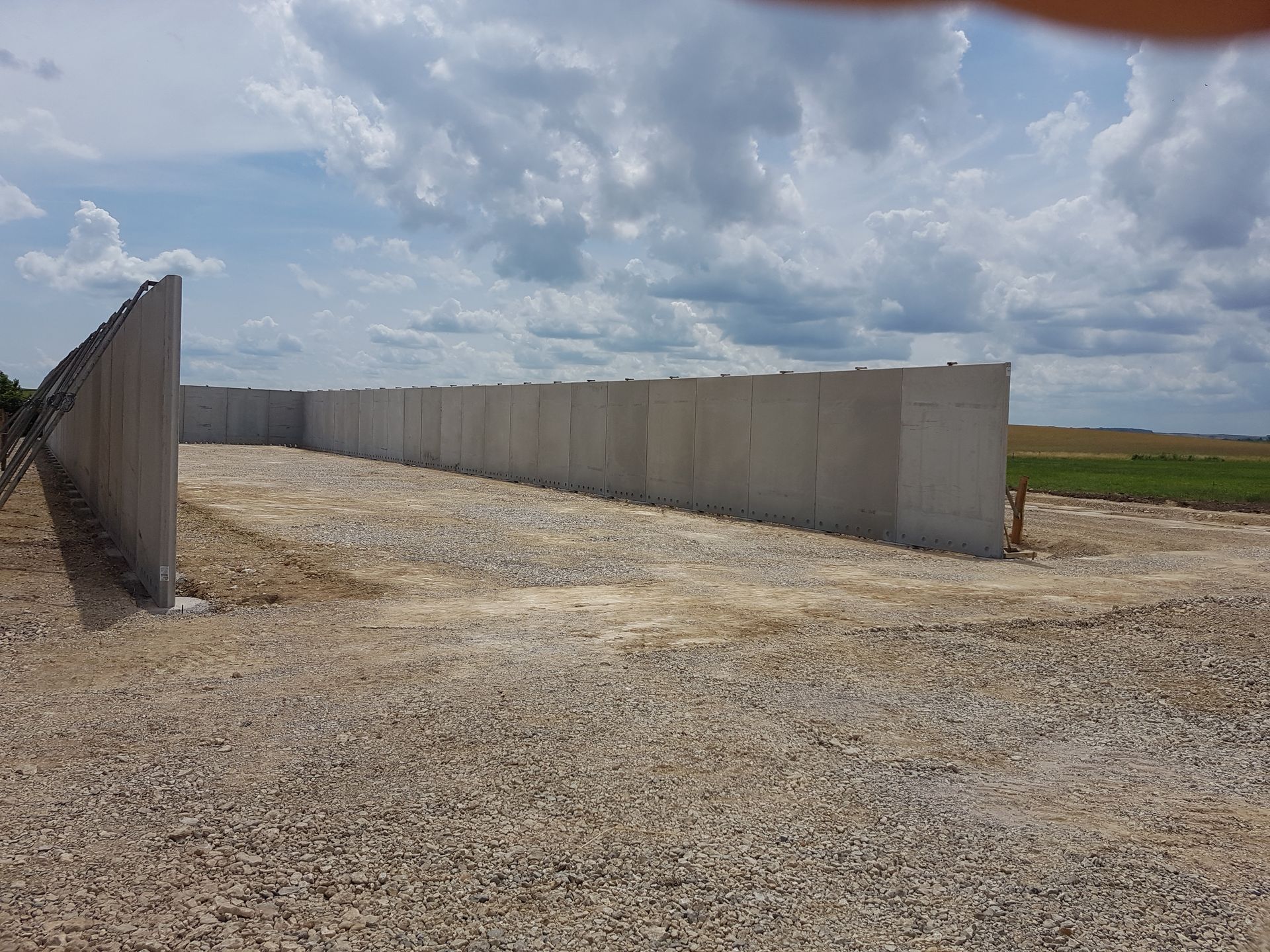 Mur en béton sur un terrain de gravier sous un ciel nuageux, à côté de champs ouverts.