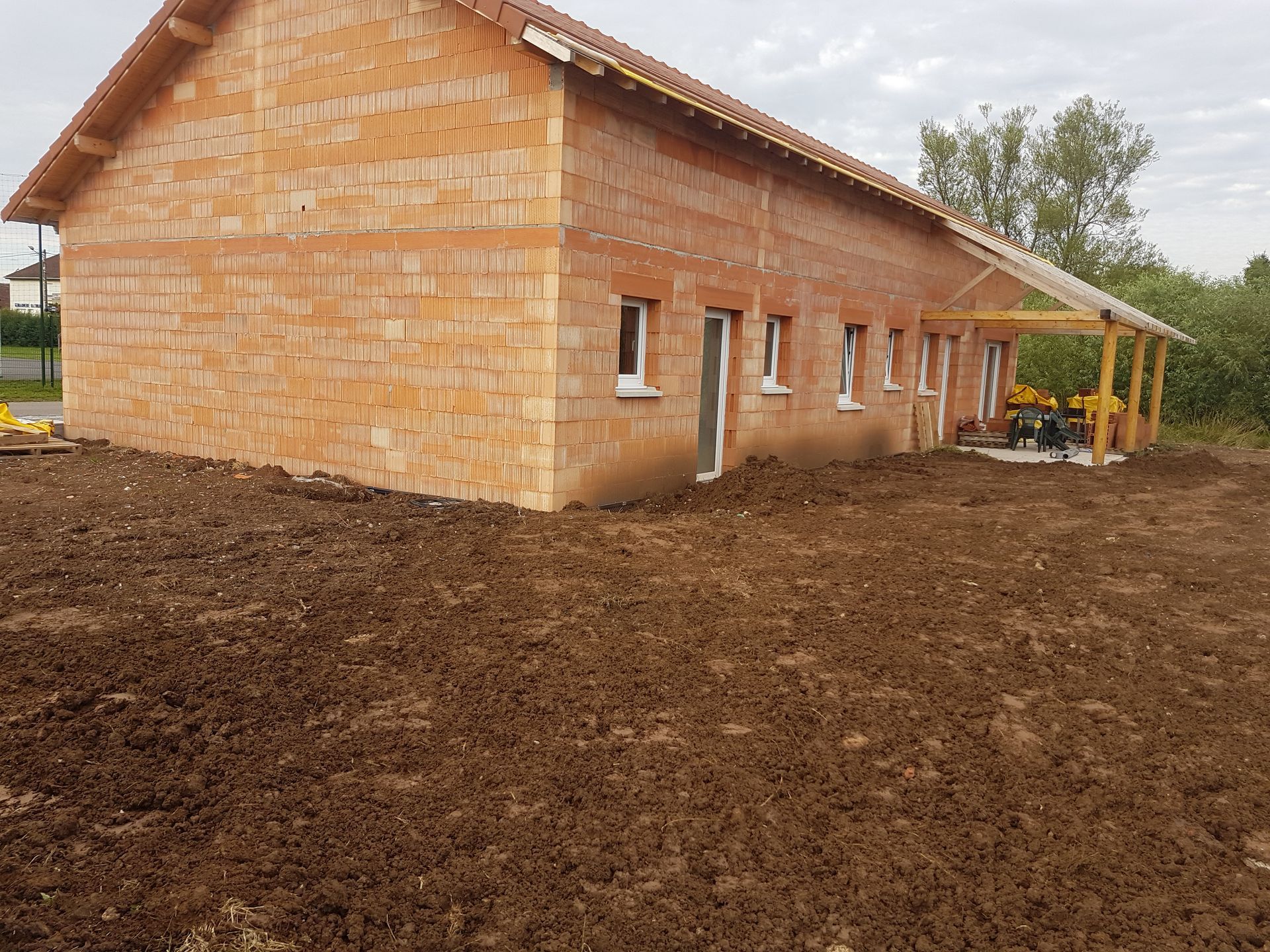Maison de plain-pied en briques en construction sur un terrain boueux, avec un porche couvert et des arbres à proximité.