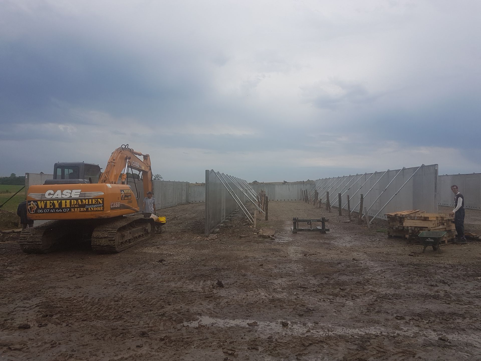 Une excavatrice et une structure de construction dans un champ boueux sous un ciel nuageux, avec des ouvriers à proximité.