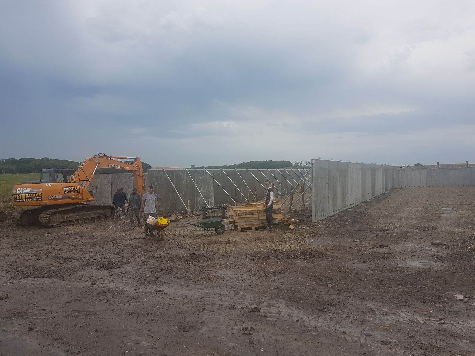 Chantier de construction avec une excavatrice à côté d'un long mur en béton sous un ciel nuageux.