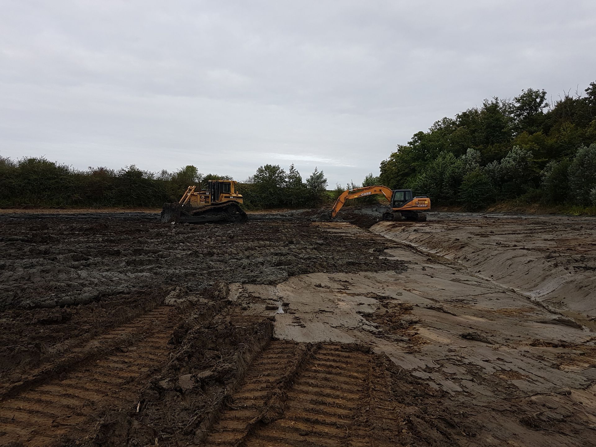 Des excavatrices travaillent sur un chantier boueux sous un ciel couvert.
