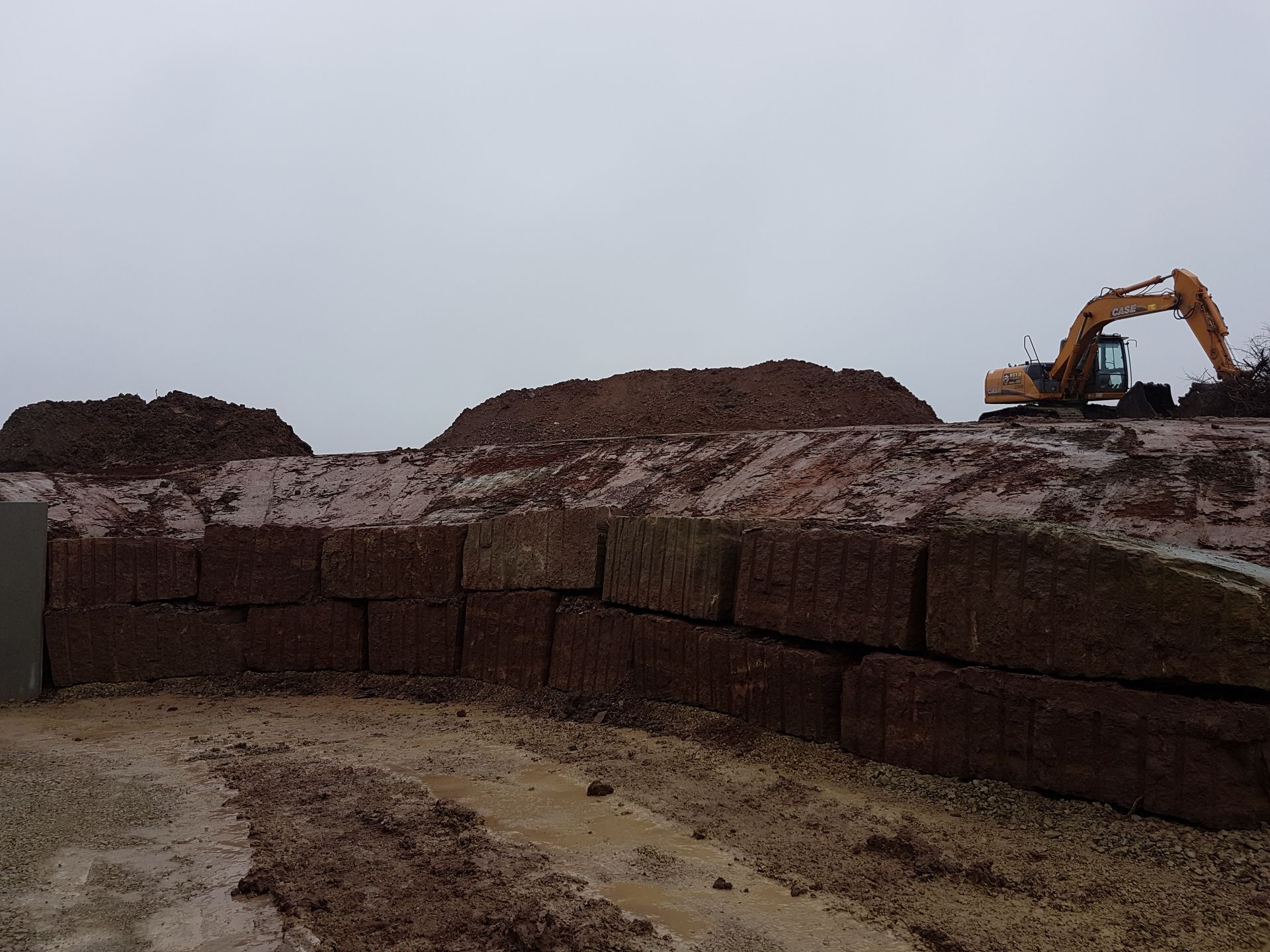 Une excavatrice travaille sur un chantier boueux, à côté d'un grand mur de terre empilée.