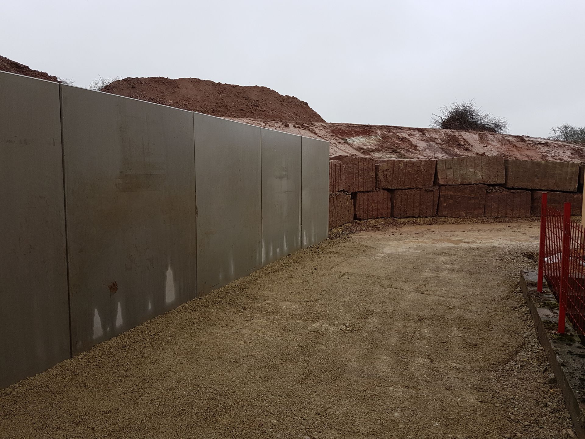 Chemin de terre longeant un haut mur gris, avec des blocs rouges empilés.