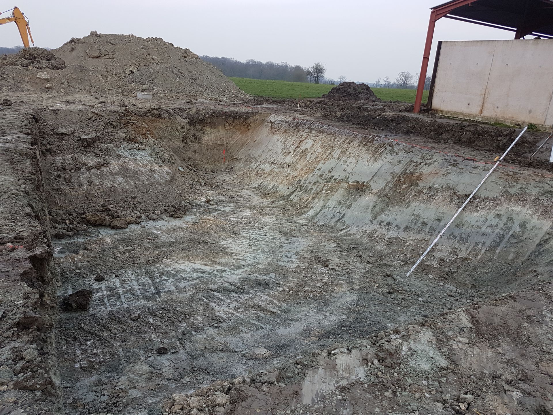 Fosse boueuse creusée à côté d'un monticule de terre et d'un bâtiment blanc sur un chantier de construction.