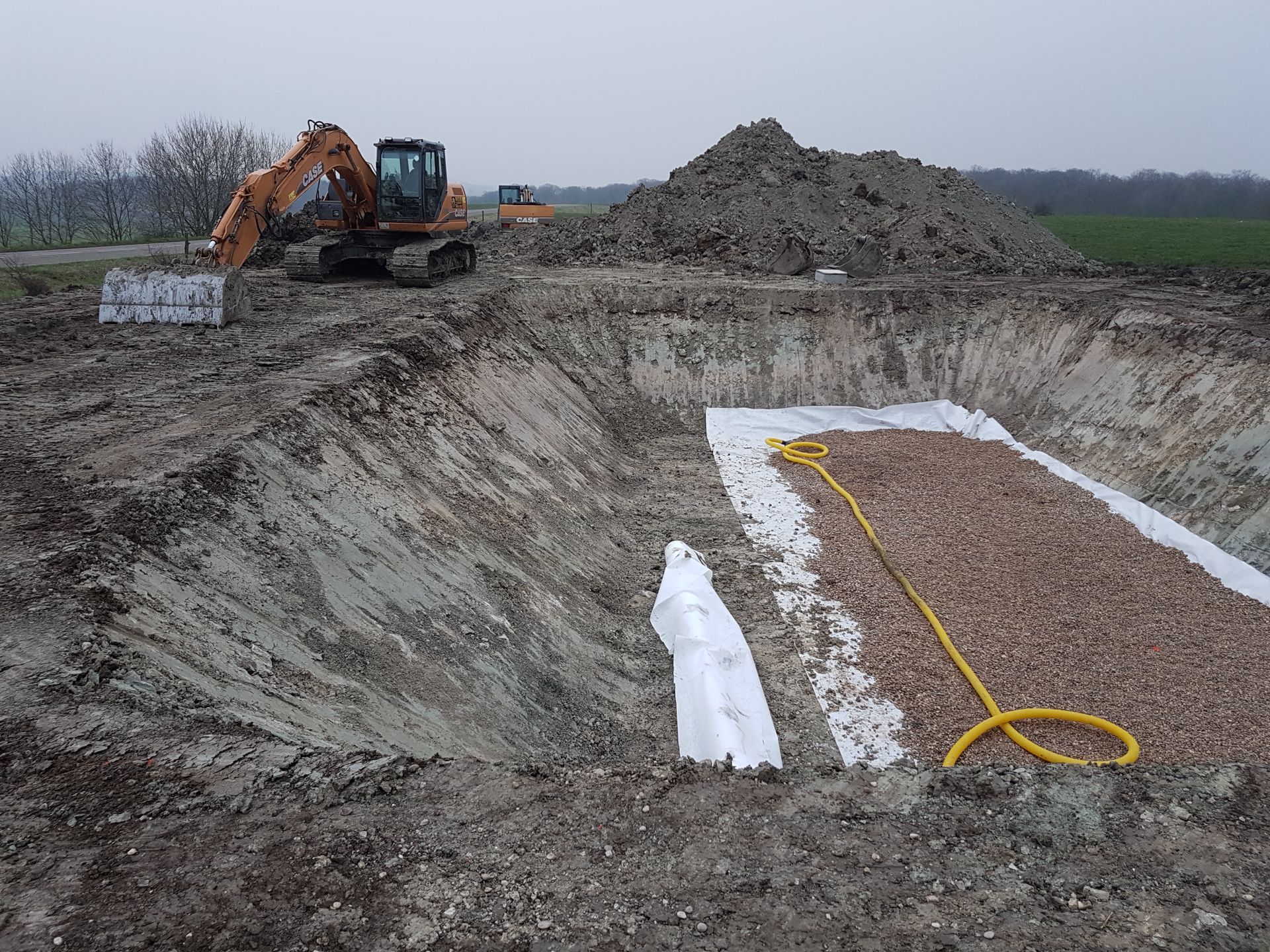 Une excavatrice sur un chantier boueux, à côté d'une tranchée recouverte d'une bâche et d'un tas de gravier.