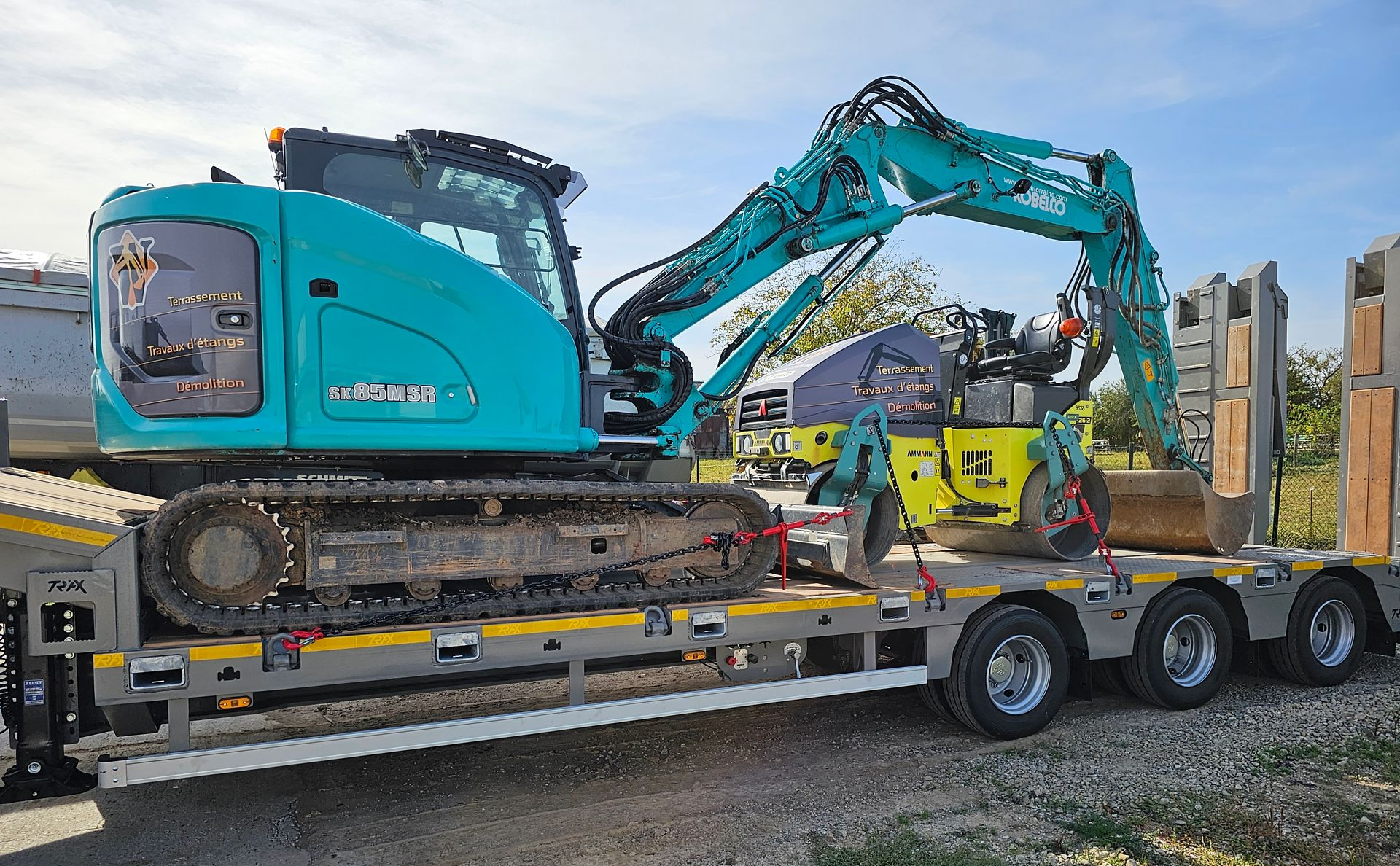 Une excavatrice turquoise est fixée sur une remorque à plateau sur un chantier de construction.